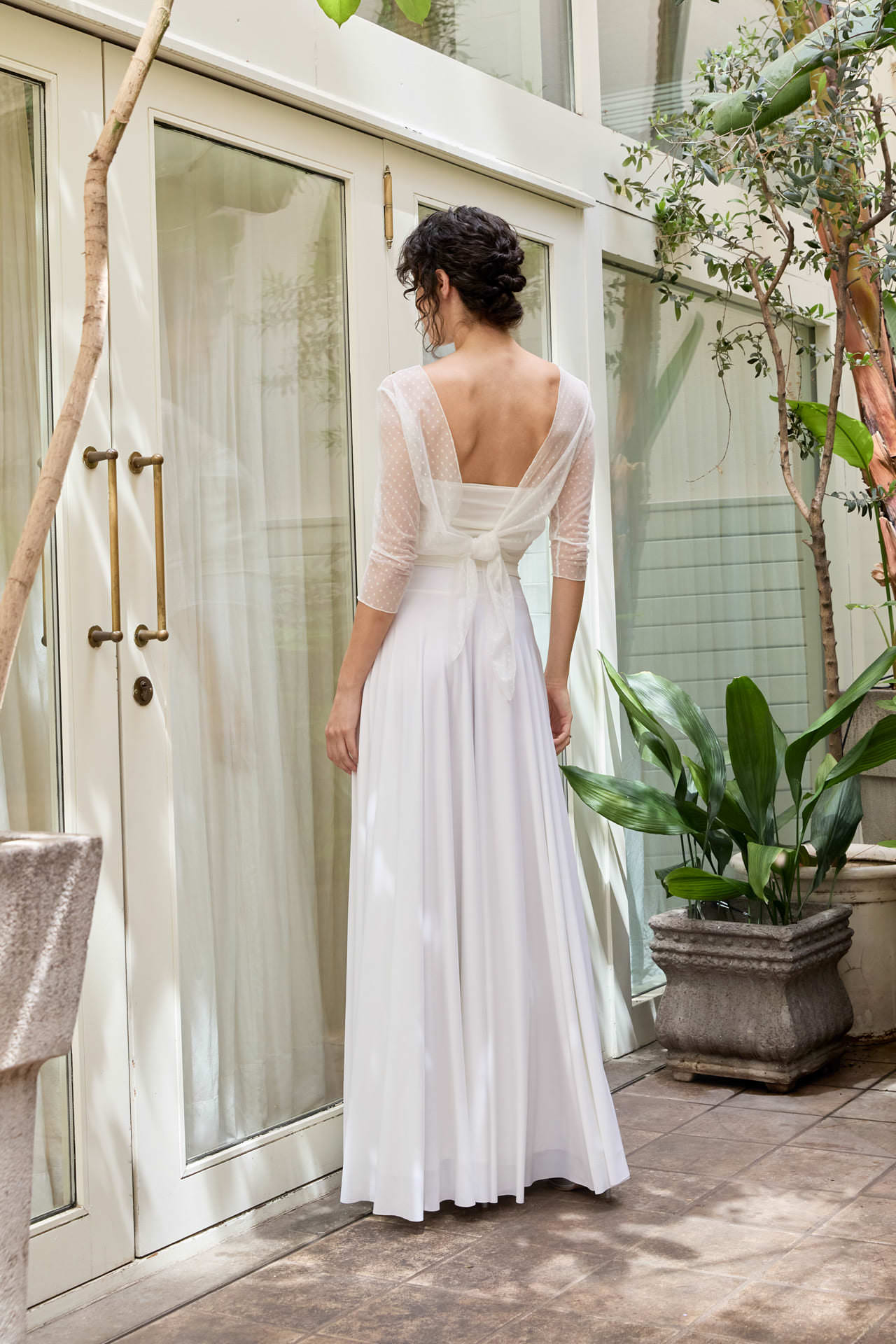 CEYLAN BCN wedding dress with an open back and sheer dotted sleeves in a glass-walled garden.